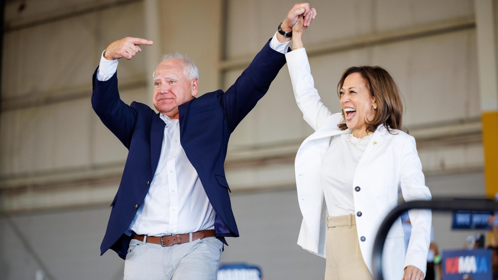 Tim Walz steht bei einer US-Wahlkampfveranstaltung neben Kamala Harris und zeigt mit Fingern auf sie, um sie ins Rampenlicht zu heben.