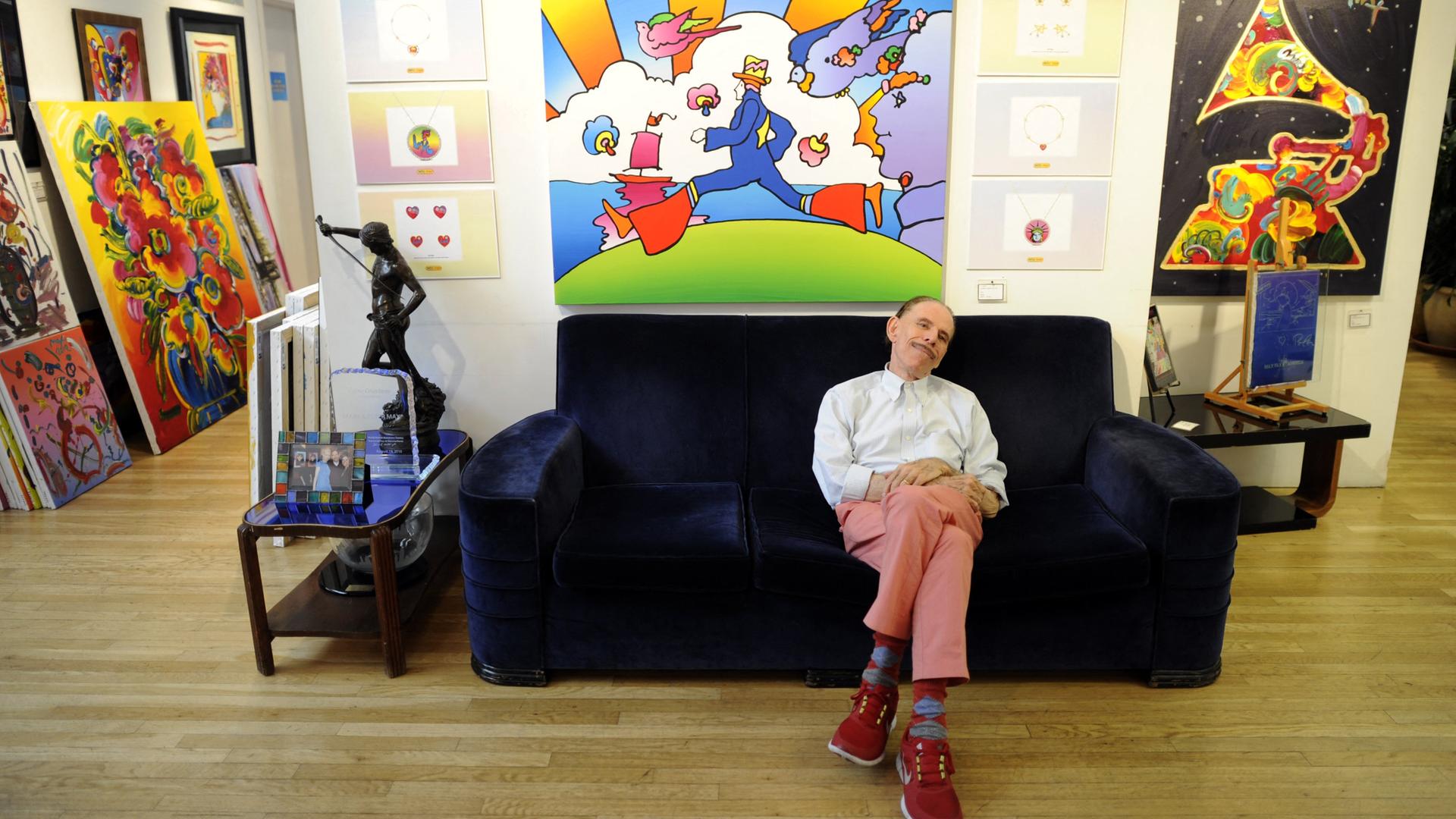 Der Pop-Art-Künstler Peter Max sitzt auf einer Couch vor einem seiner Bilder.
