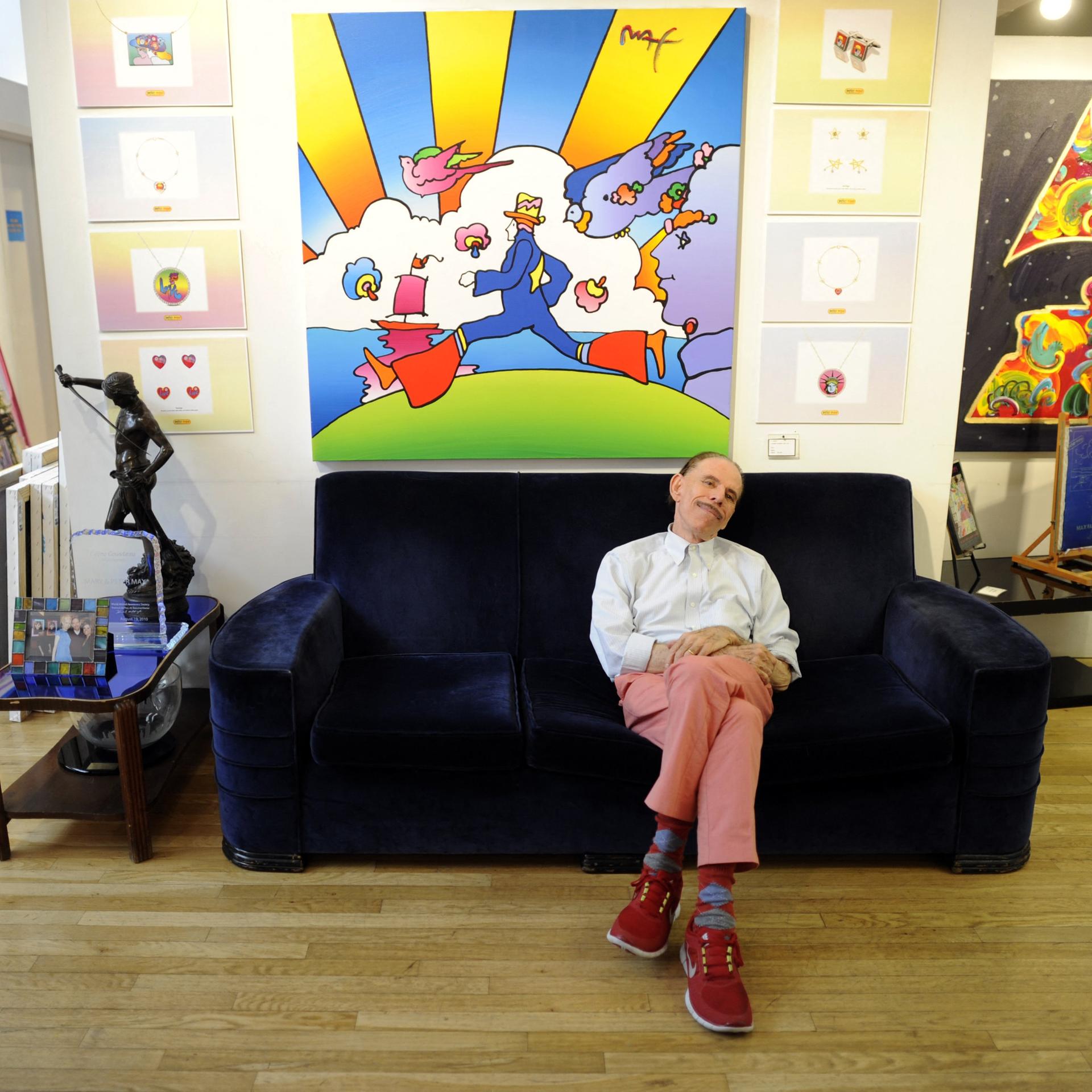 Der Pop-Art-Künstler Peter Max sitzt auf einer Couch vor einem seiner Bilder.