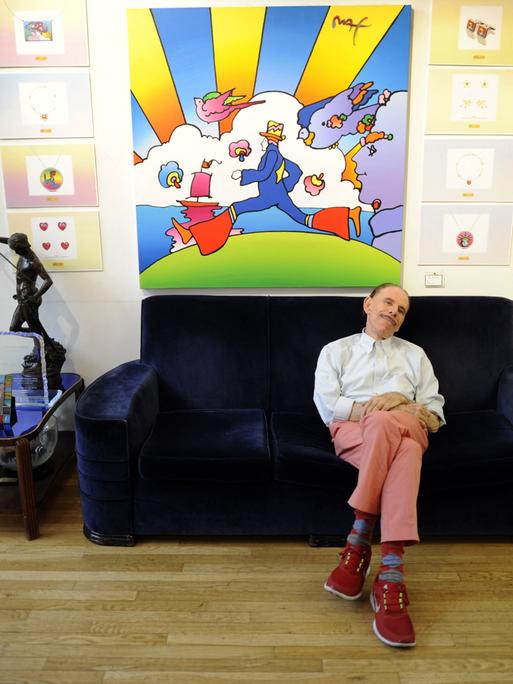 Der Pop-Art-Künstler Peter Max sitzt auf einer Couch vor einem seiner Bilder. Der Pop-Art-Künstler Peter Max sitzt auf einer Couch vor einem seiner Bilder.