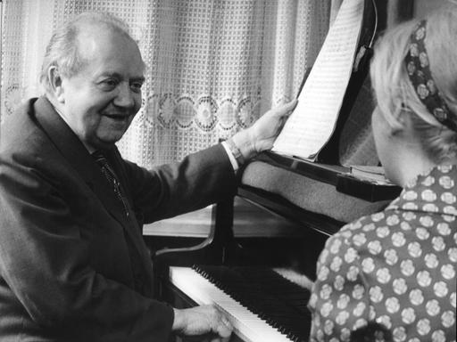 Auf dem schwarz-weiß Bild ist ein älterer Mann am Klavier zu sehen, der einer Hand Noten hält und eine Frau anlächelt, die von hinten zu sehen ist.
