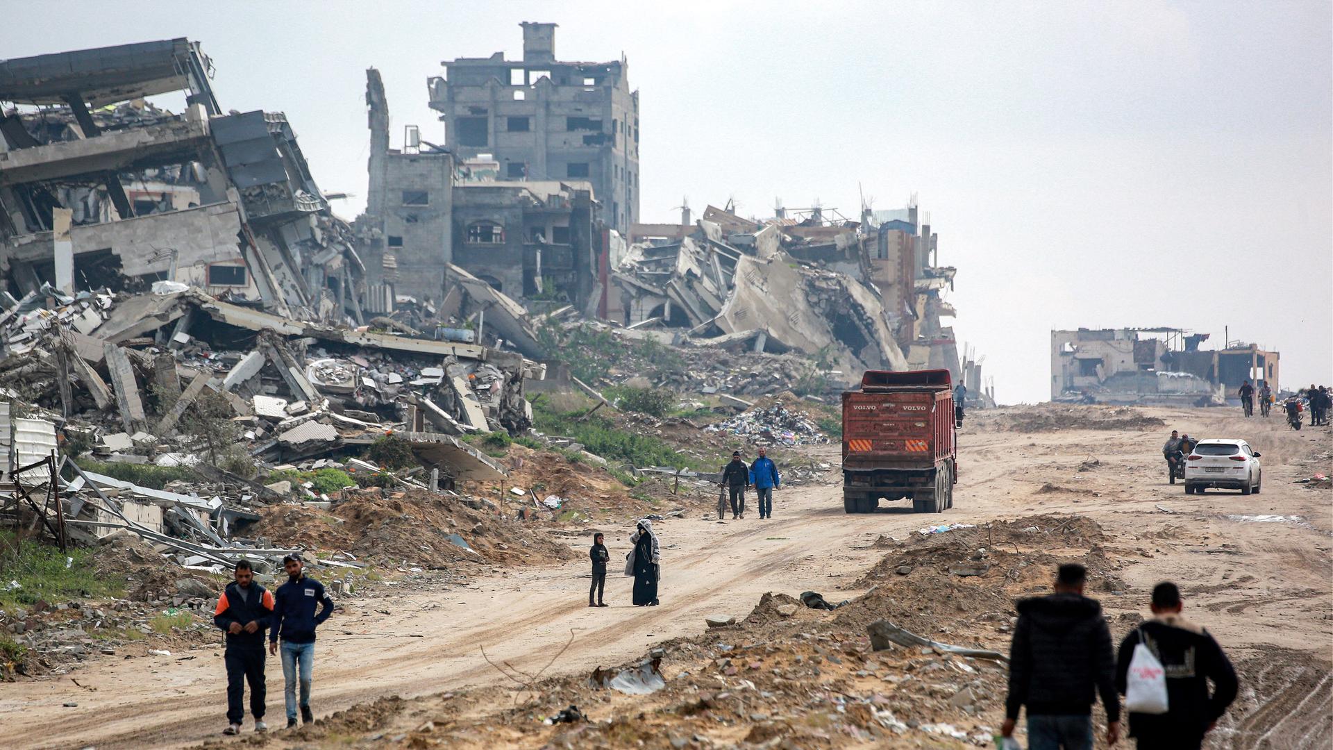 Menschen gehen auf einer unbefestigten Straße im Gazastreifen an völlig zerstörten Gebäuden vorbei.