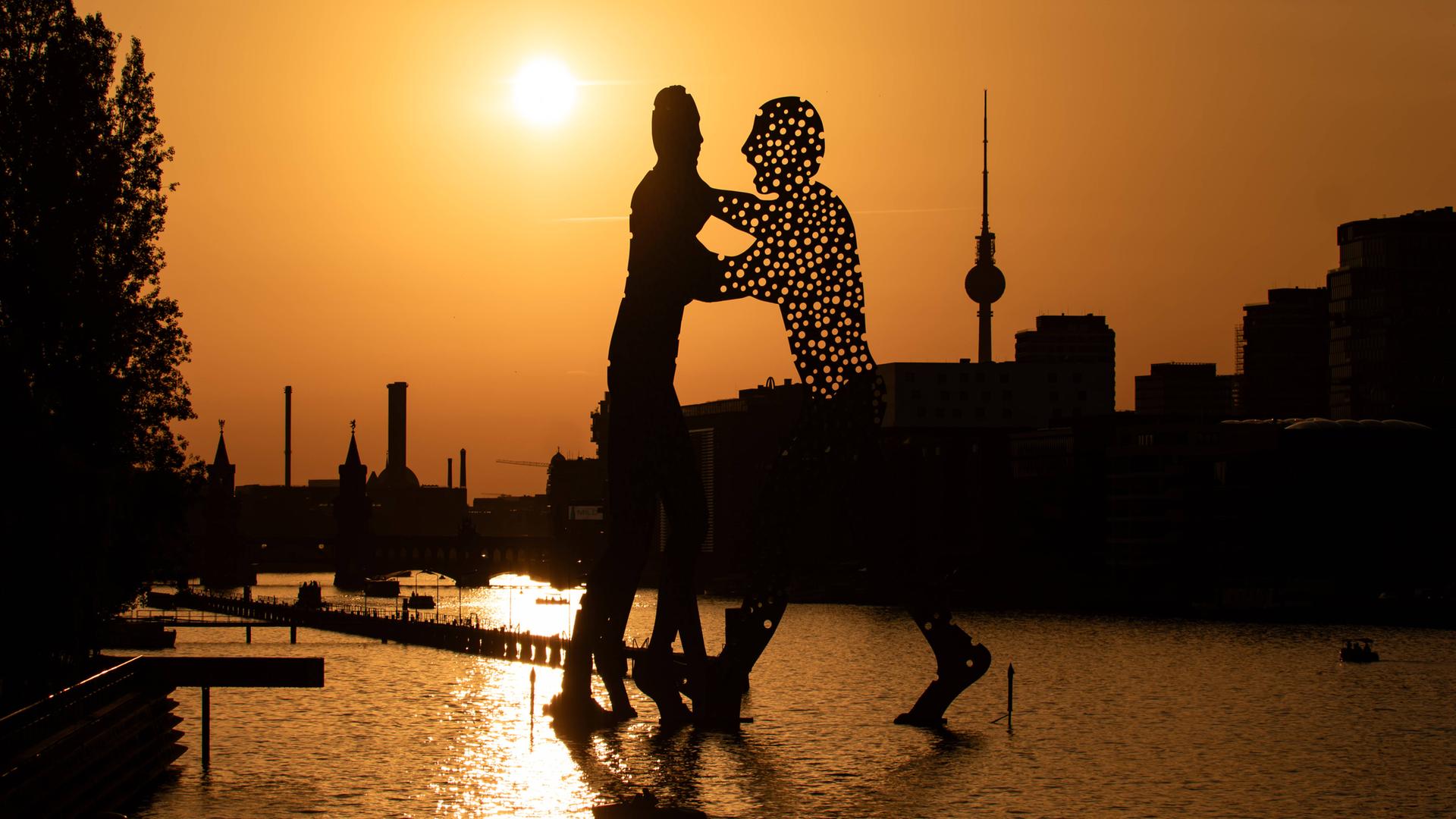 Eine rötliche Abendsonne scheint hinter der Skulptur Molecular Men an der Spree in Berlin am 24. Juno 2026, die eine scharze Silhouette von zwei Menschen darstellt. Dahinter die Berliner Skyline mit Fernsehturm