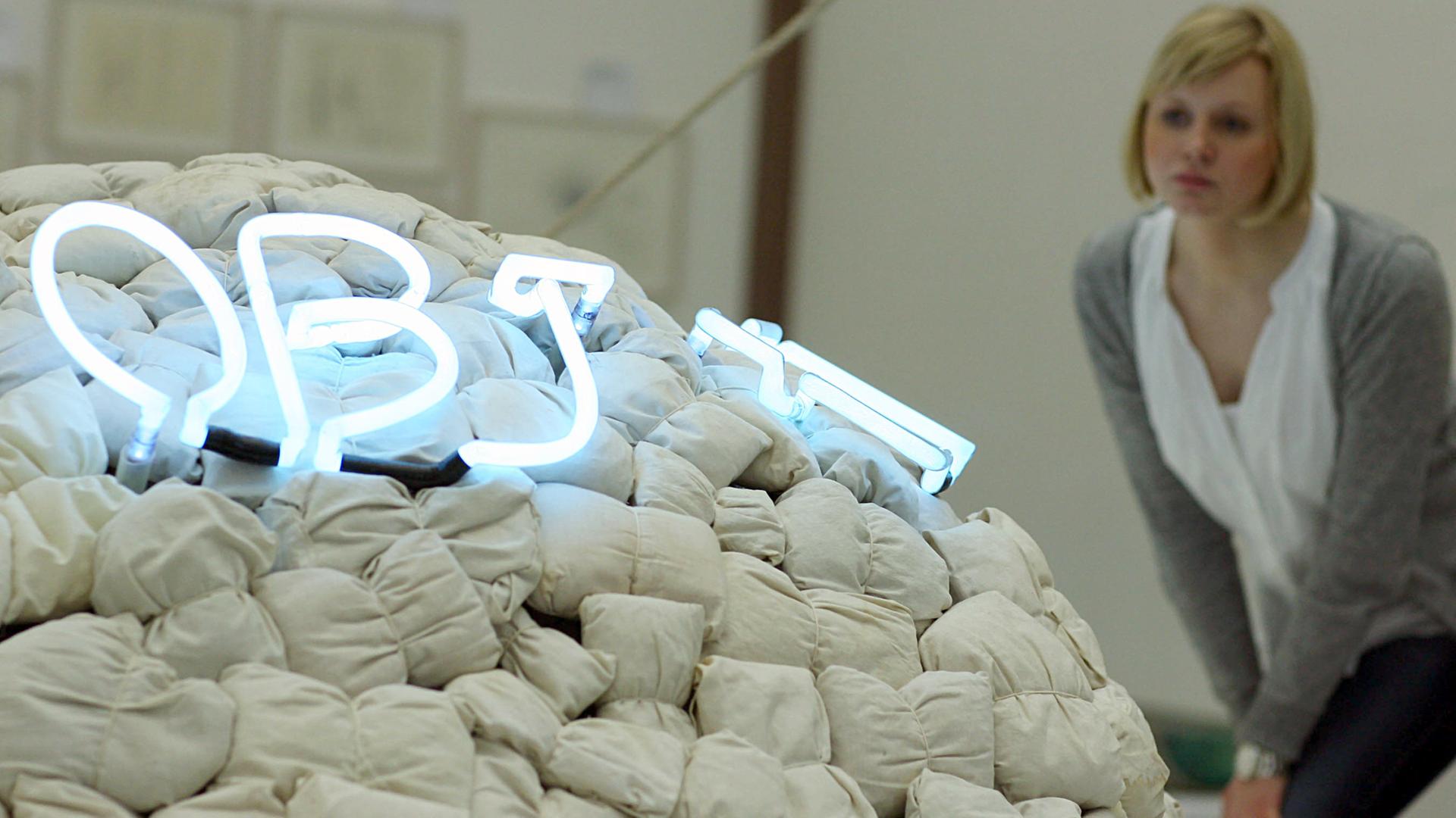 Eine Frau schaut auf ein Kunstwerk von Mario Merz mit dem Titel "Objet cache-toi" (Objekt versteck dich). Mit Holzwolle gefüllte Leichensäcke, die zu einem Iglu geformt sind. 