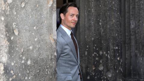 Tom Schilling in einem grauen Anzug an einer grauen Mauer