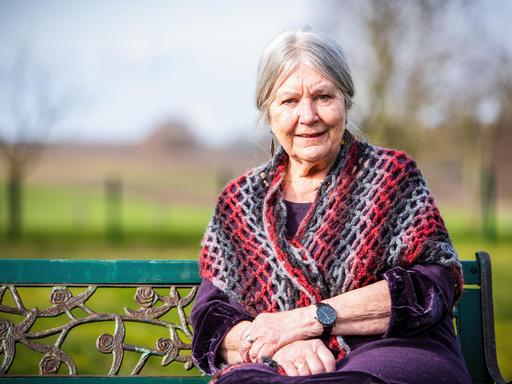 Helga Schubert hat graune Haare und trägt eine bunte Weste. Sie sitzt auf einer Bank umgeben von Natur.