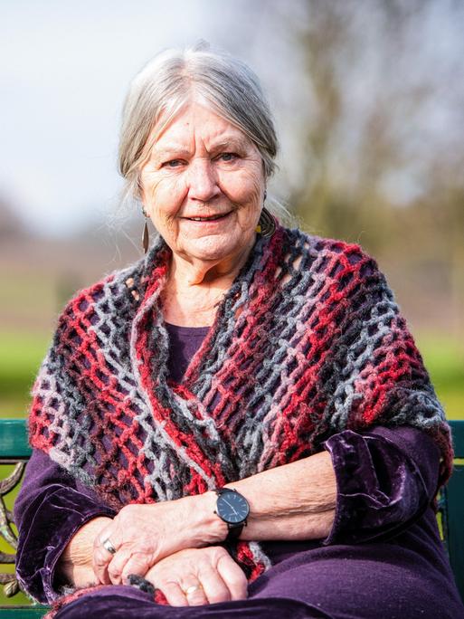 Helga Schubert hat graune Haare und trägt eine bunte Weste. Sie sitzt auf einer Bank umgeben von Natur.