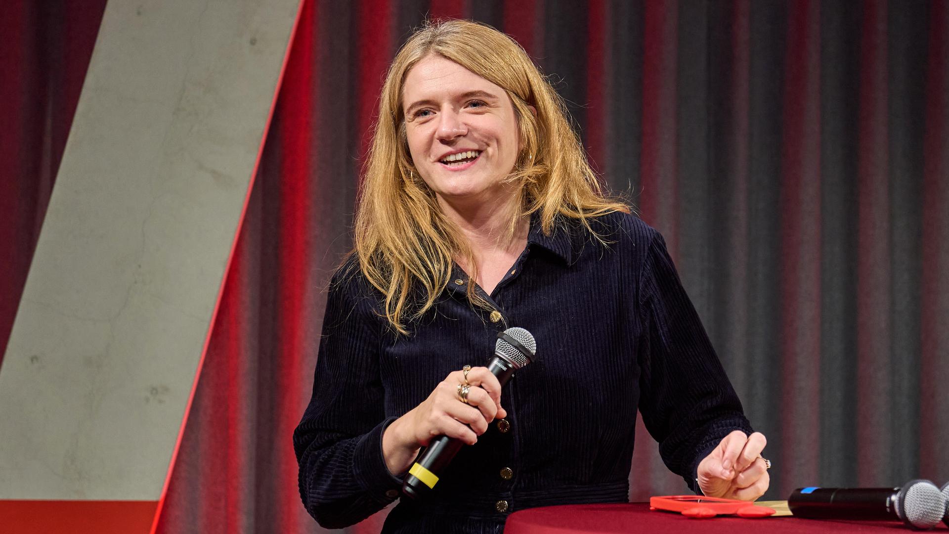 Katrin Gottschalk steht mit einem Mikrofon in der Hand an einem Tisch. Katrin Gottschalk steht mit einem Mikrofon in der Hand an einem Tisch.