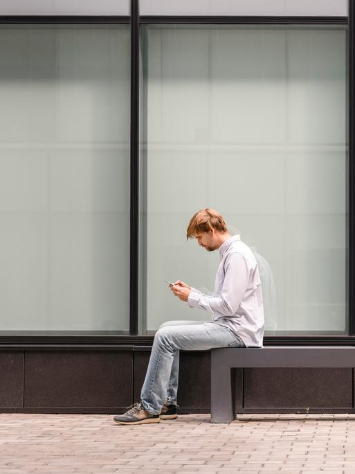 Zwei Männer sitzen mit Abstand auf einer Bank und schauen auf ihr Handy.