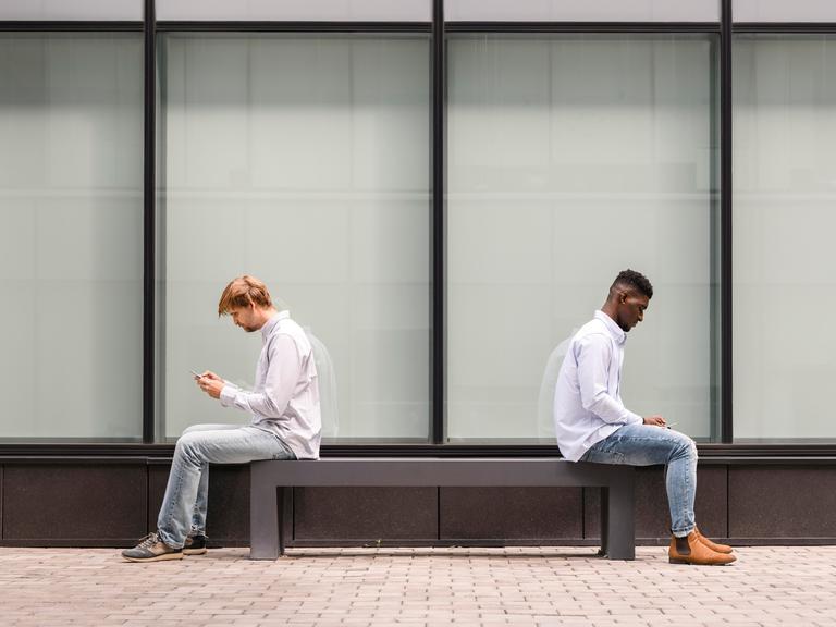 Zwei Männer sitzen mit Abstand auf einer Bank und schauen auf ihr Handy.