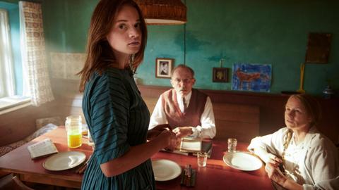 Im Filmstill aus "Was man von hier aus sehen kann" steht Luna Wedler im grünen Kleid in einer Küche und schaut direkt in die Kamera. Hinter ihr am Esstisch sitzen ihre Großeltern und schauen zu ihr auf.