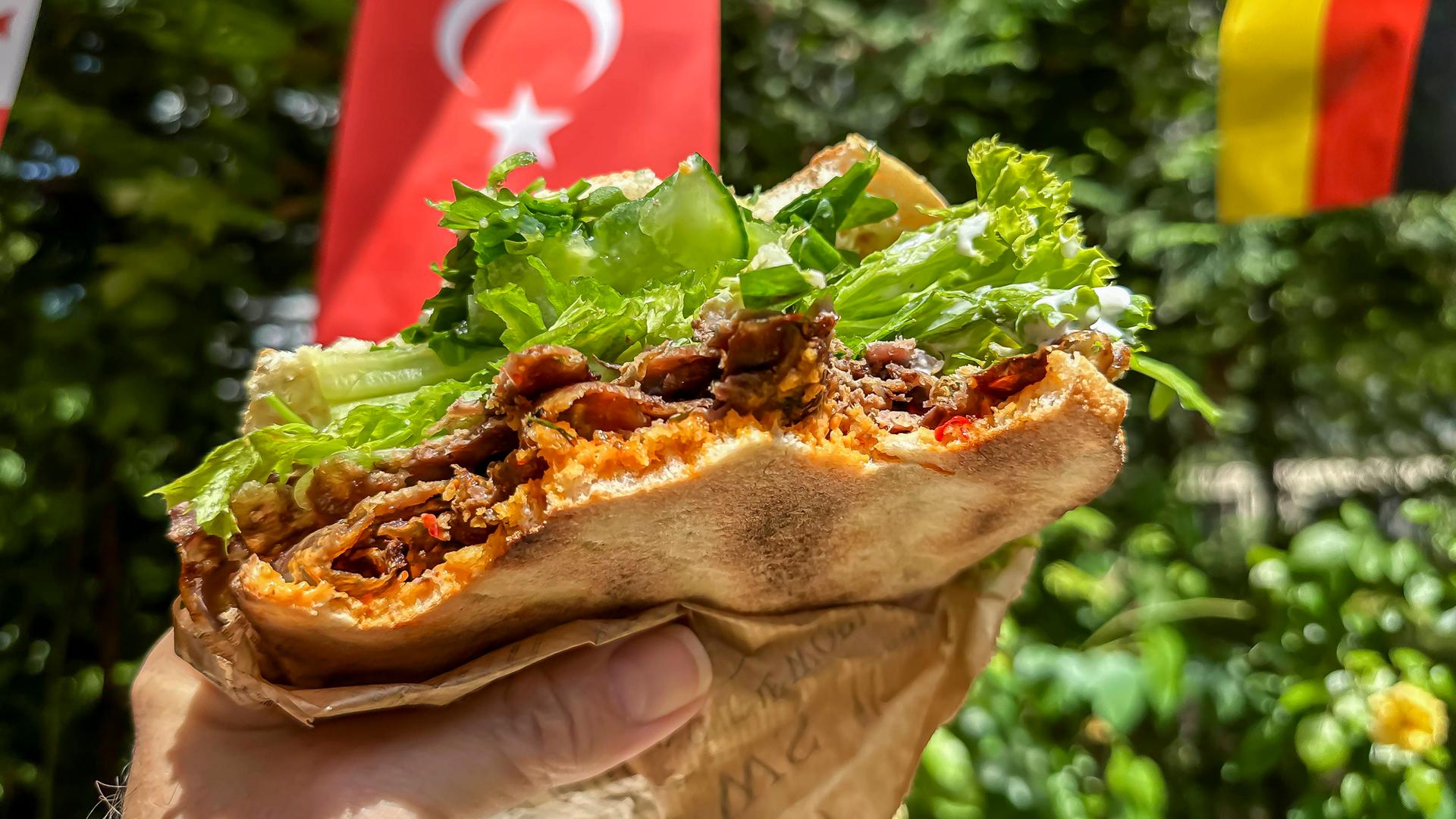 Ein Mann hält einen Döner-Kebab-Sandwich in einer Hand in die Kamera. Im Hintergrund sind türkische und deutsche Fähnchen zu sehen.