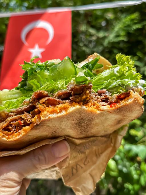 Ein Mann hält einen Döner-Kebab-Sandwich in einer Hand in die Kamera. Im Hintergrund sind türkische und deutsche Fähnchen zu sehen.