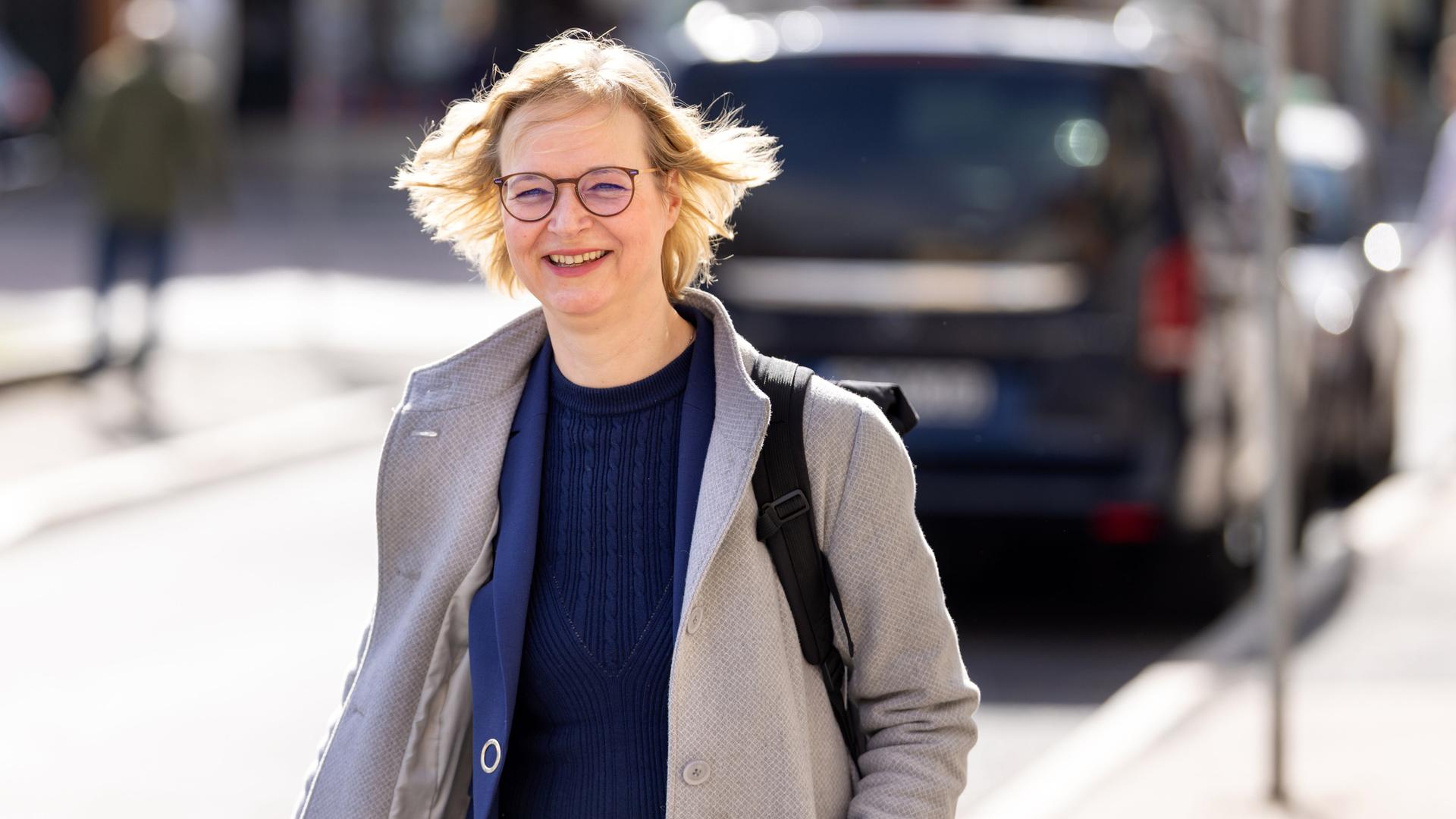 Katja Wolf, Oberbürgermeisterin von Eisenach, läuft mit schwingendem Mantel über die Straße und lächelt in die Kamera.