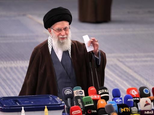 Irans oberster Führer Ajatollah Chamenei bei der Abgabe seiner Stimme. Vor einer Wahlurne hält er einen weißen Zettel in der Hand, vor ihm zahlreiche Mikrofone
