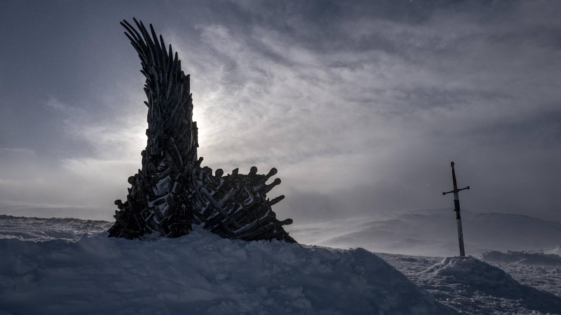 In einer düsteren Schneelandschaft steht der Thron aus der Fantasy-Serie Game of Thrones, daneben steckt ein Schwert im Schnee.