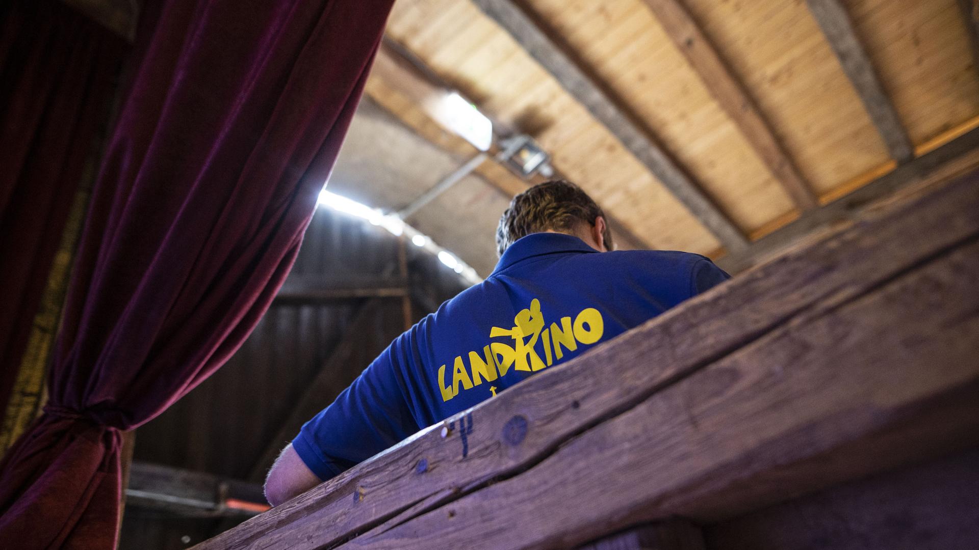 In einer Scheune sitzt ein Mann auf einer Bank und trägt ein blaues Shirt mit der Aufschrift "Landkino" und einem Kinoprojektor.