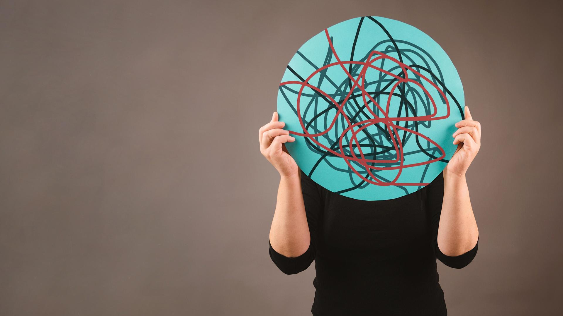 Eine Person hält sich ein rundes Schild vor das Gesicht, auf dem chaotisch verlaufende Linien zu sehen sind