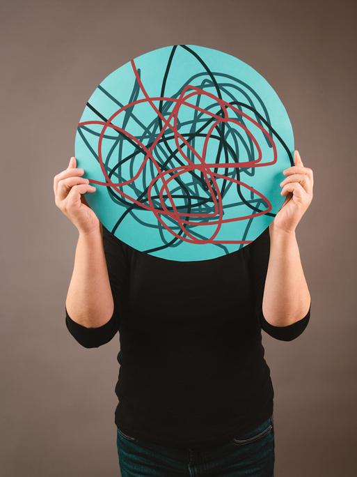 Eine Person hält sich ein rundes Schild vor das Gesicht, auf dem chaotisch verlaufende Linien zu sehen sind Eine Person hält sich ein rundes Schild vor das Gesicht, auf dem chaotisch verlaufende Linien zu sehen sind