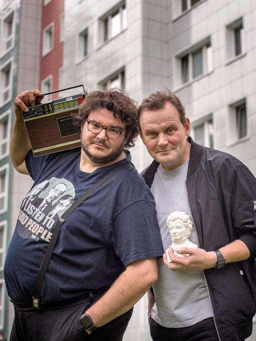 Devid Striesow und Axel Ranisch stehen vor einem Plattenbau. Devid Striesow hält den Kopf einer kleinen Statue auf dem Arm, Axel Ranisch ein Radio auf der Schulter.