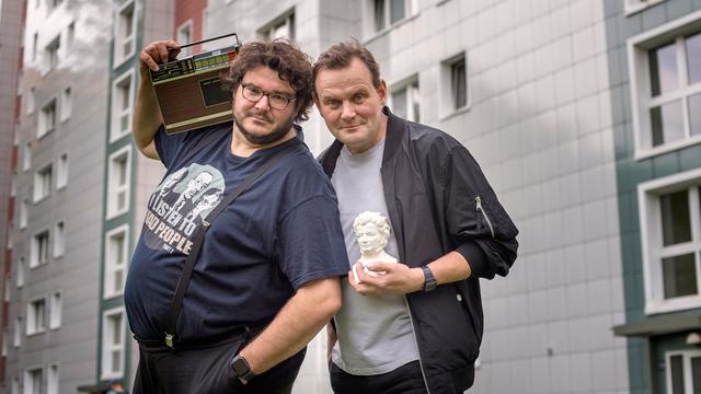 Devid Striesow und Axel Ranisch stehen vor einem Plattenbau. Devid Striesow hält den Kopf einer kleinen Statue auf dem Arm, Axel Ranisch ein Radio auf der Schulter.