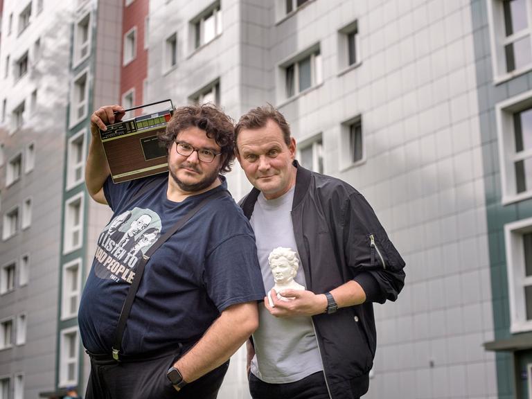 Devid Striesow und Axel Ranisch stehen vor einem Plattenbau. Devid Striesow hält den Kopf einer kleinen Statue auf dem Arm, Axel Ranisch ein Radio auf der Schulter.