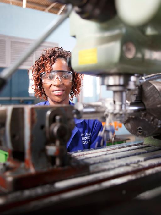 Eine Frau steht hinter einer Maschine, trägt eine Schutzbrille und lächelt in die Kamera. 