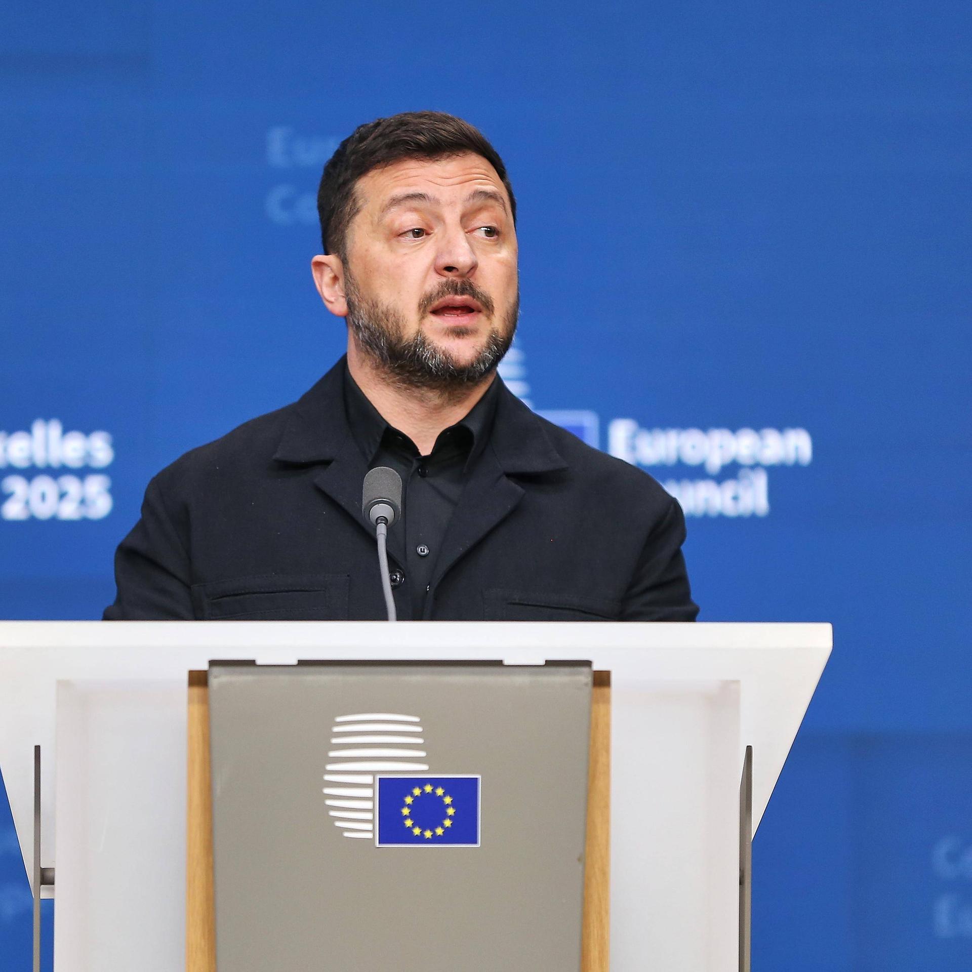 Der ukranische Präsident Selenskyj gibt beim EU-Gipfel in Brüssel eine Pressekonferenz. Er steht an einem Rednerpult und trägt ein schwarzes Hemd sowie ein schwarzes Sakko. Hinter ihm eine blaue Wand mit dem Logo des Gipfels in weißer Schrift. Der ukranische Präsident Selenskyj gibt beim EU-Gipfel in Brüssel eine Pressekonferenz. Er steht an einem Rednerpult und trägt ein schwarzes Hemd sowie ein schwarzes Sakko. Hinter ihm eine blaue Wand mit dem Logo des Gipfels in weißer Schrift.