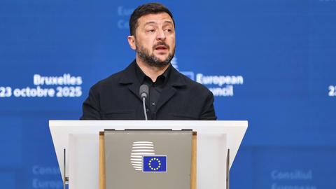 Der ukranische Präsident Selenskyj gibt beim EU-Gipfel in Brüssel eine Pressekonferenz. Er steht an einem Rednerpult und trägt ein schwarzes Hemd sowie ein schwarzes Sakko. Hinter ihm eine blaue Wand mit dem Logo des Gipfels in weißer Schrift. 
