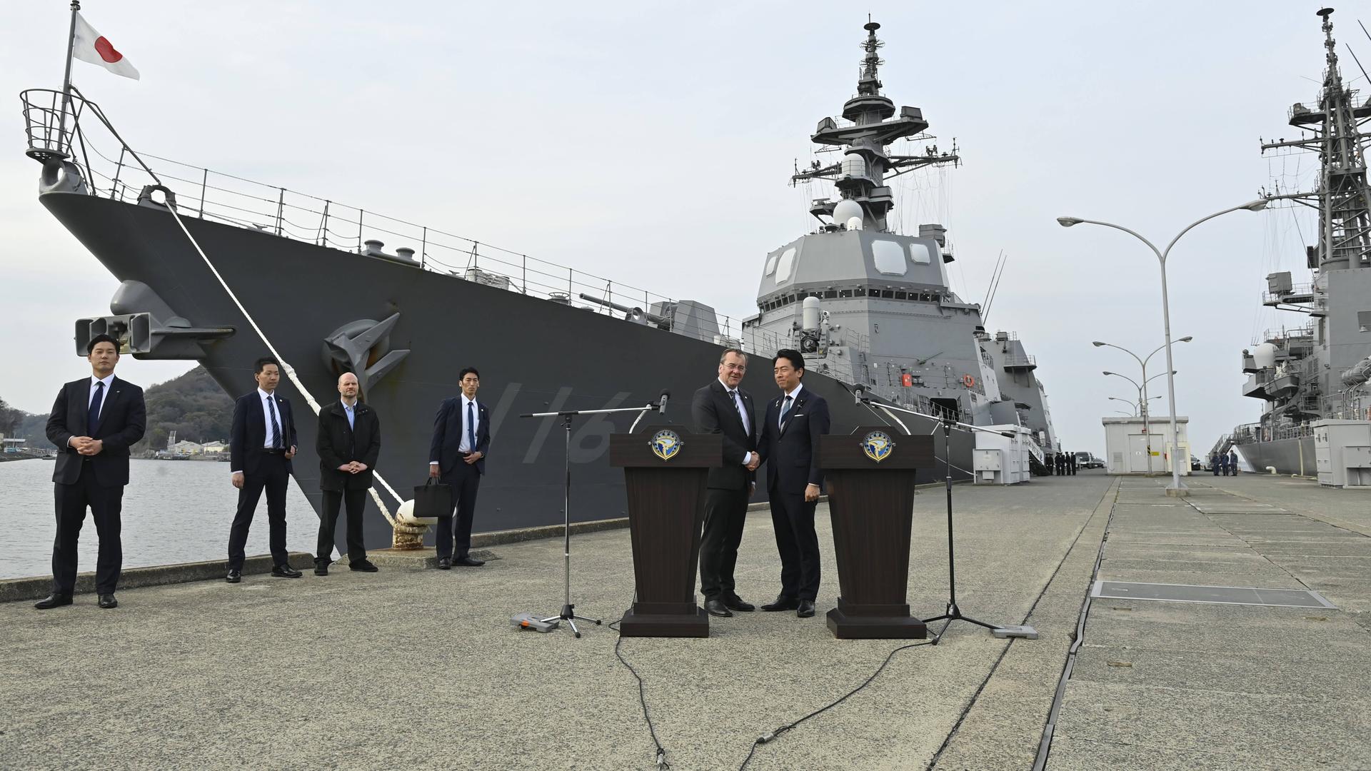 Pistorius und Koizumi schütteln an zwei Rednerpulten sich die Hand. Sie stehen in einem Hafen, im Hintergrund ist ein japanisches Kriegsschiff zu sehen. Pistorius und Koizumi schütteln an zwei Rednerpulten sich die Hand. Sie stehen in einem Hafen, im Hintergrund ist ein japanisches Kriegsschiff zu sehen.