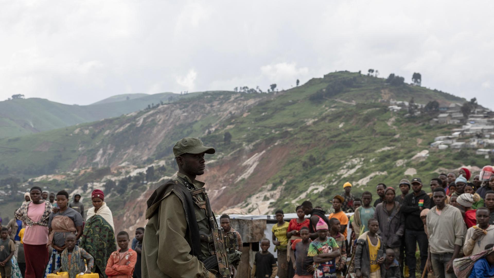 Ein Milizionär der M23-Rebellengruppe steht an den Coltan-Gruben in Rubaya. Im Hintergrund stehen Menschen. Am Horizont sind die Lagerstätten von Coltan-Erz, das im Tagebau gefördert wird. 