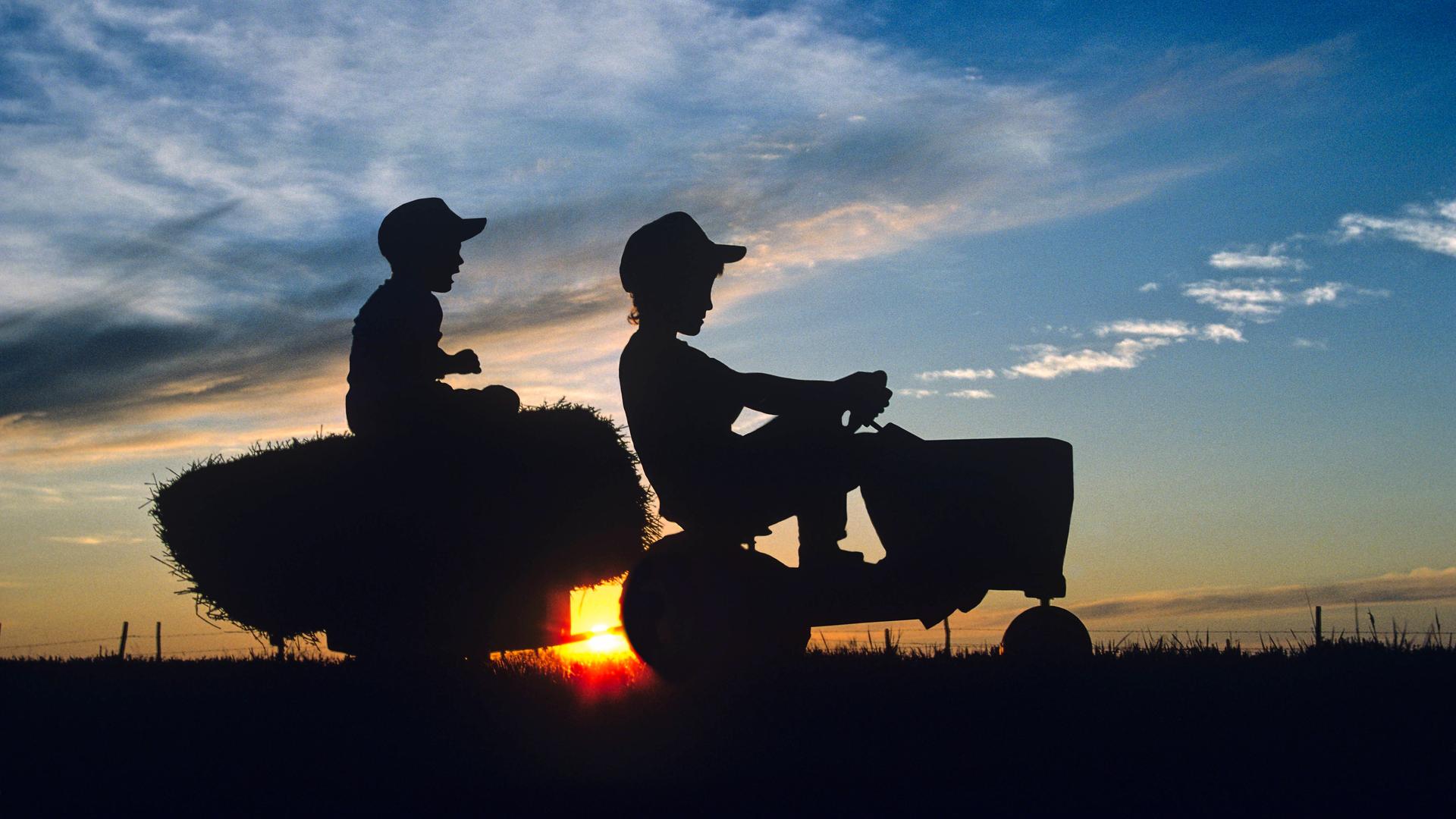 2 Jungen auf einem Trecker mit Anhänger auf einem Feld bei Sonnenuntergang