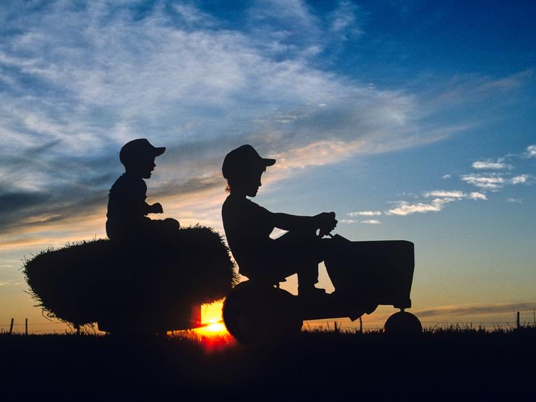 2 Jungen auf einem Trecker mit Anhänger auf einem Feld bei Sonnenuntergang