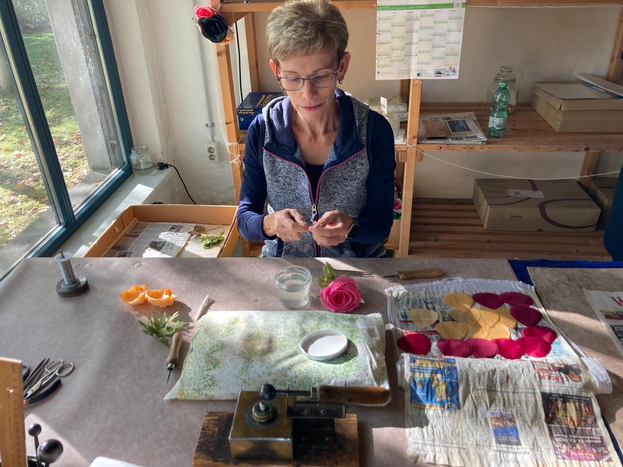 Eine Mitarbeiterin der Kunstblumenmanufaktur Heide Steyer sitzt an ihrem Arbeitstisch und fertigt Blüten aus Stoff. Vor ihr auf dem Tisch liegt ein Steckkissen, daneben Blütenblätter aus Stoff sowie bereits fertiggestellte Blumen. Die Frau trägt eine Brille und hat kurze graue Haare.