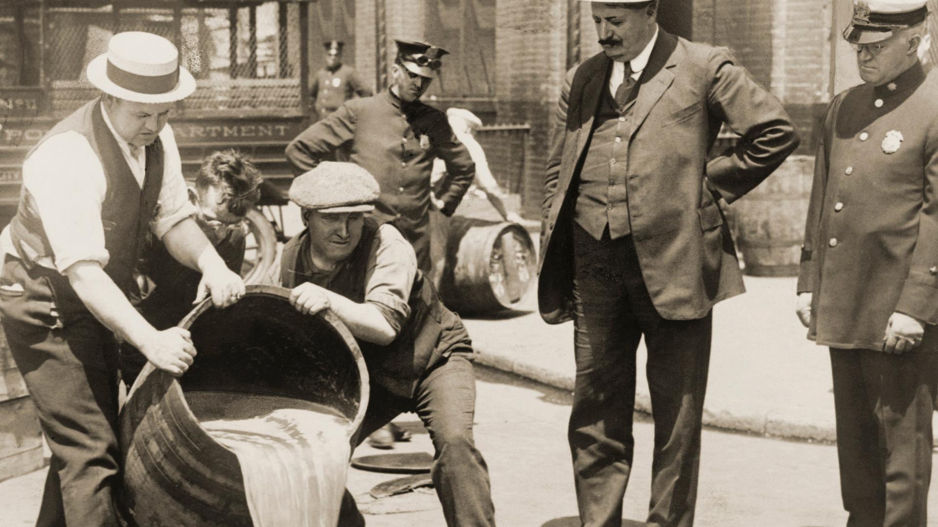 Ein Polizist und andere Männer schauen Anfang der 1920er-Jahre in New York dabei zu, wie zwei Personen ein Fass in einen Gulli entleeren.