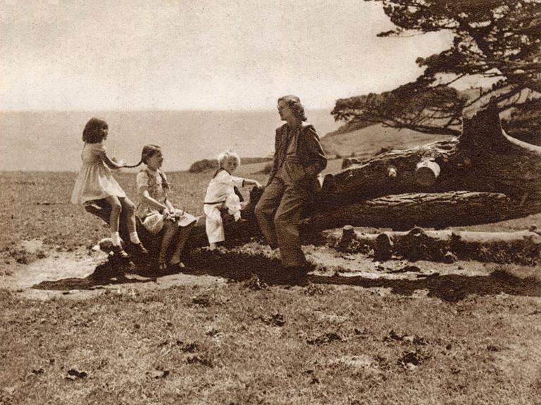 Daphne du Maurier sitzt mit ihren Kindern am Strand von Cornwall auf einem umgefallenen Baum. Im Hintergrund ist das Meer zu sehen.