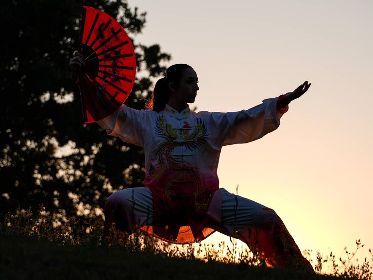 Silhouette einer Tai-Chi-Athletin mit Fächer und vor Sonnenuntergang Silhouette einer Tai-Chi-Athletin mit Fächer und vor Sonnenuntergang