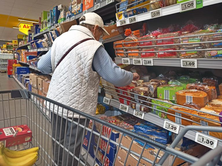 Eine alte Frau nimmt in einem Supermarkt Brot aus einem Regal.  
