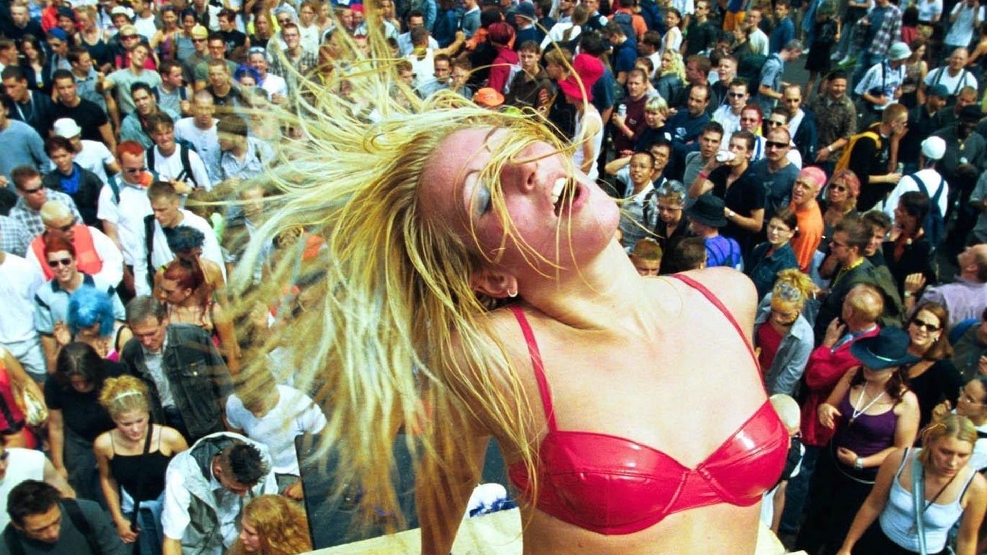 Eine blonde Frau im pinken Bikini und mit Plateauschuhen tanzt auf einem Wagen der Love Parade vor hunderten Menschen.