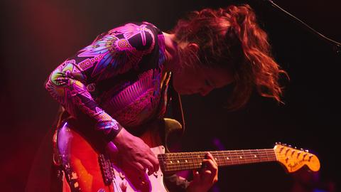Eine Frau beugt sich in bunter Kleidung und bunt beleuchtet über eine E-Gitarre.