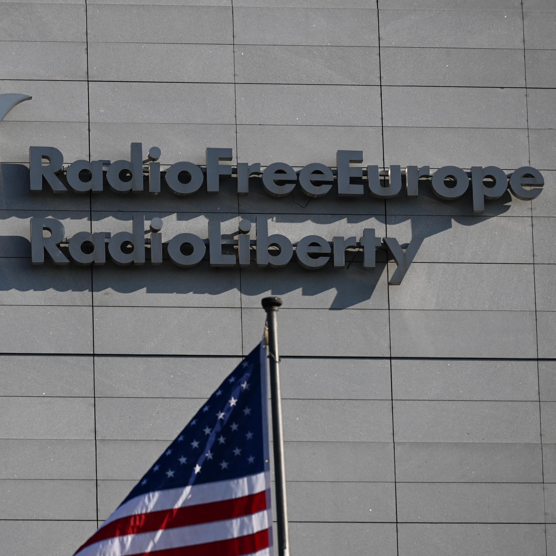 An einer Wand steht der Schriftzug "Radio Free Europe/Radio Liberty" und das Logo des Senders. Davor steht eine US-Flagge. 