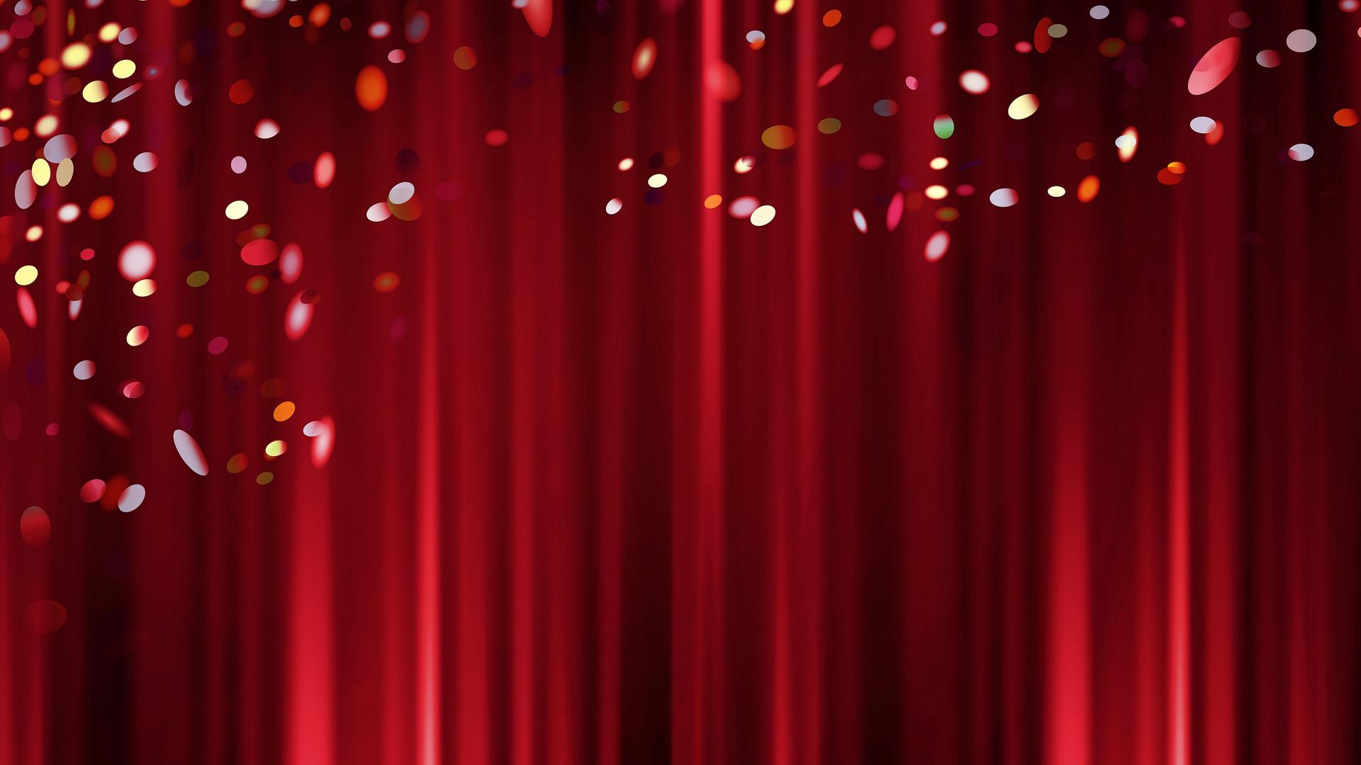 Das Bild zeigt einen dunkelroten, geschlossenen Theatervorhang. Im Vordergrund rieselt buntes Konfetti von der Decke. 