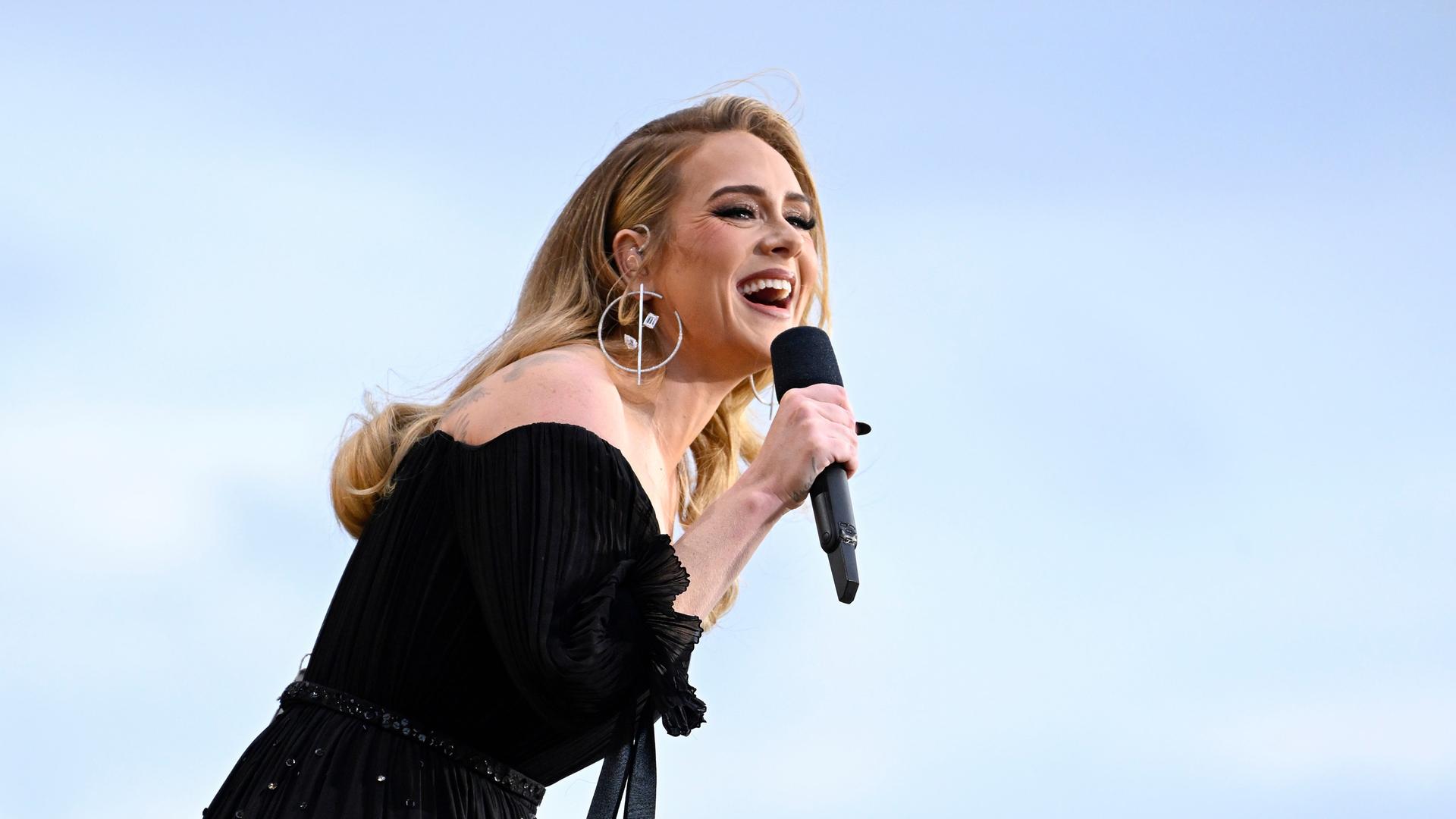 Die Sängerin Adele auf der Bühne in schwarzem Kleid vor blauem Himmel mit einem Mikrofon in der Hand.