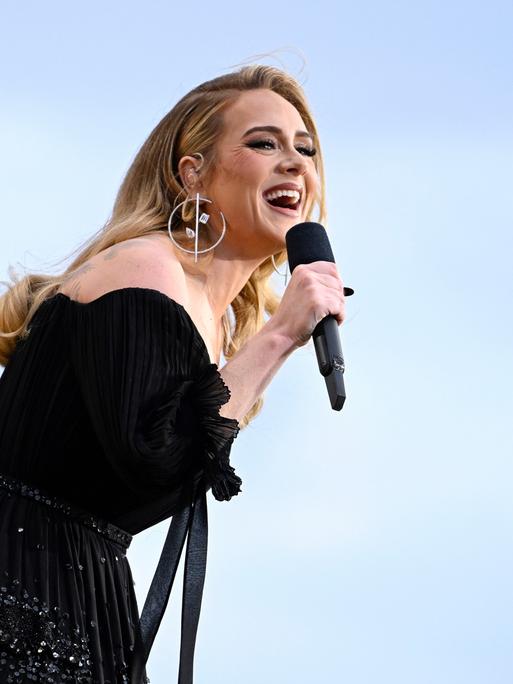 Die Sängerin Adele auf der Bühne in schwarzem Kleid vor blauem Himmel mit einem Mikrofon in der Hand.