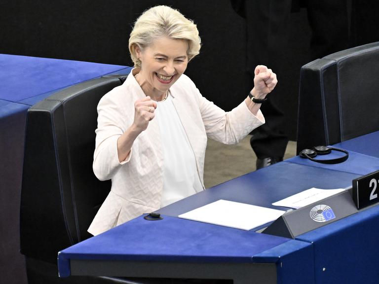 Ursula von der Leyen sitzt im Europäischen Parlament, schmeißt die Hände in die Luft und grinst.