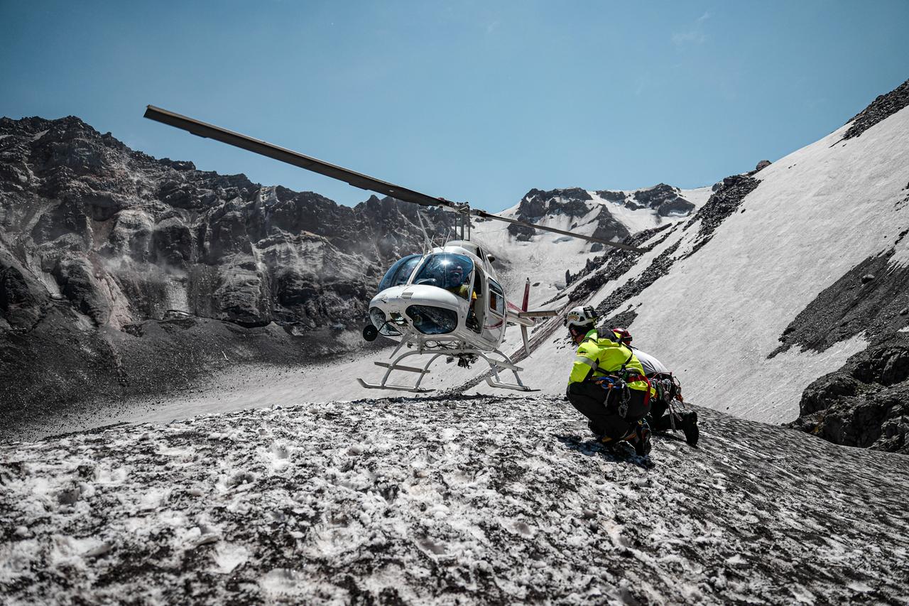 Helikopter landet in schneebedeckter Berglandschaft. Menschen stehen daneben.