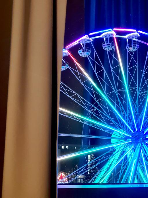 Durch ein Fenster ist ein erleuchtetes Riesenrad zu sehen. Durch ein Fenster ist ein erleuchtetes Riesenrad zu sehen.