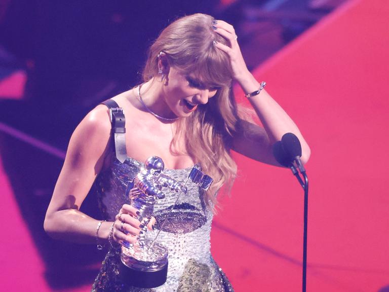 Taylor Swift steht auf der Bühne und hält in ihrer Hand den MTV Music Video Award.Eine Hand fährt durch ihr Haar, sie schaut nach unten und lacht dabei. 