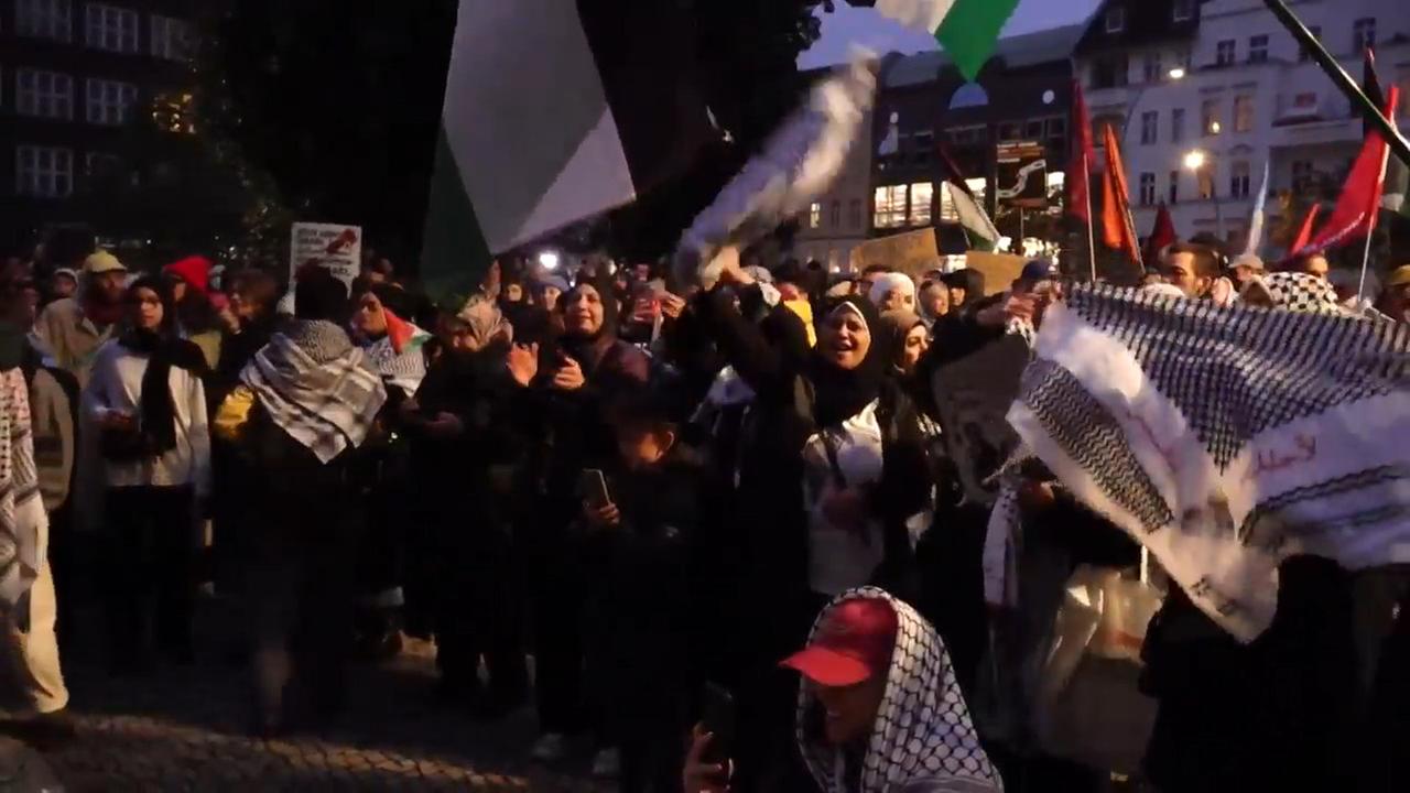 Es ist dunkel: die Demonstrantengruppe ist nur schwach beleuchtet. Die Teilnehmer tragen Fahnen, klatschen in die Hände und skandieren etwas.