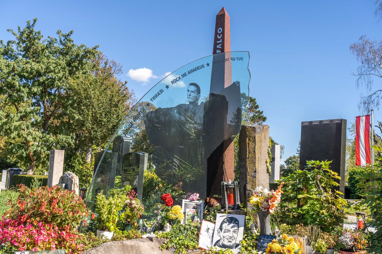 Blick auf das Grab des Sängers „Falco“ auf dem Wiener Zentralfriedhof. Auf einer Glasplatte ist der Musiker in einem schwarzen Umhang dargestellt. Darüber sind Titel seiner Songs aufgeführt, z.B. „Out oft the dark“, „Rock me Amadeus“ und „Jeanny“